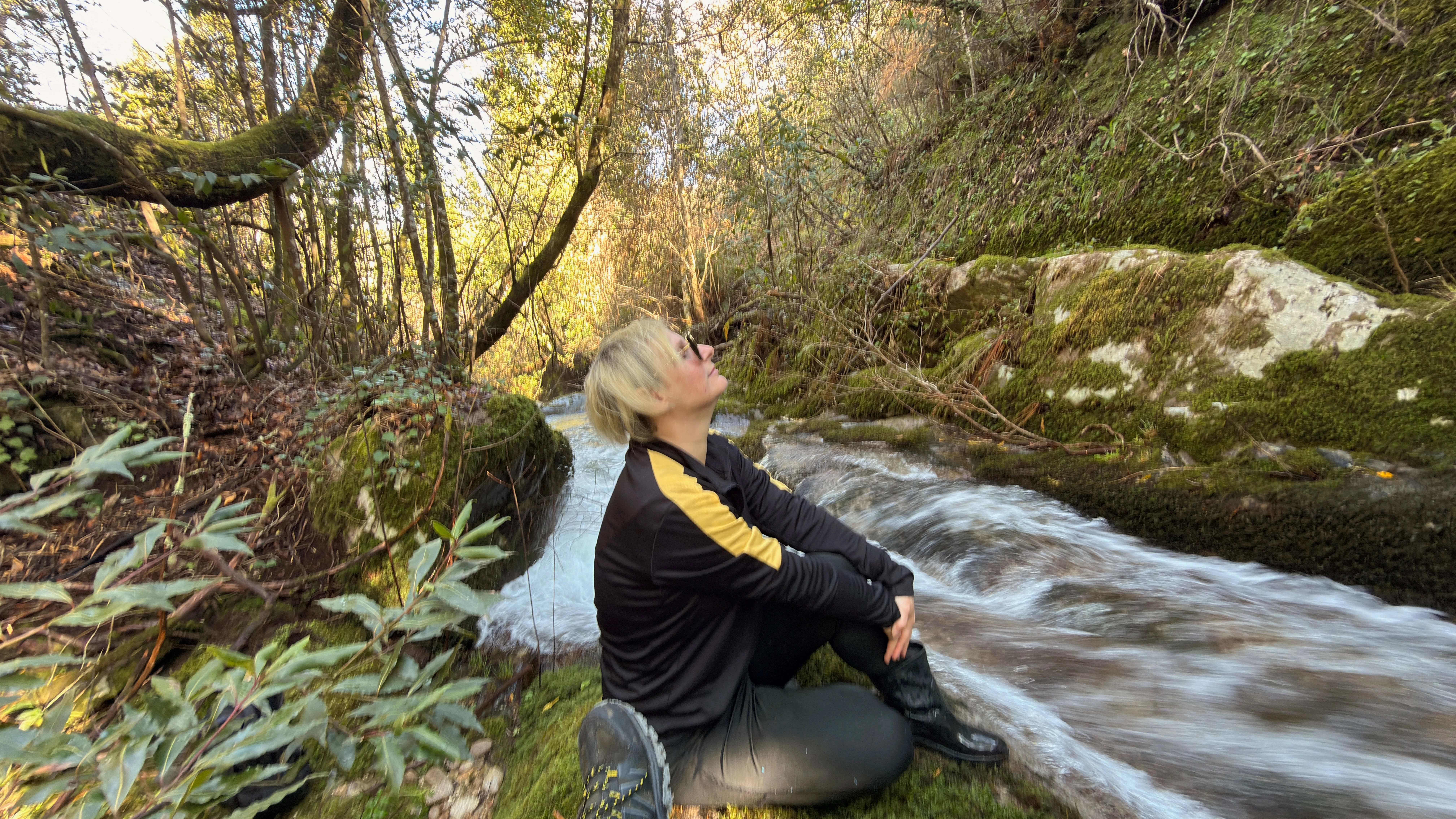 Pessoa sentada junto ao rio entre pedras e vegetação.